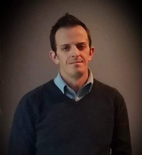 A professional portrait of a man wearing a dark sweater and light blue shirt, standing against a neutral background.
